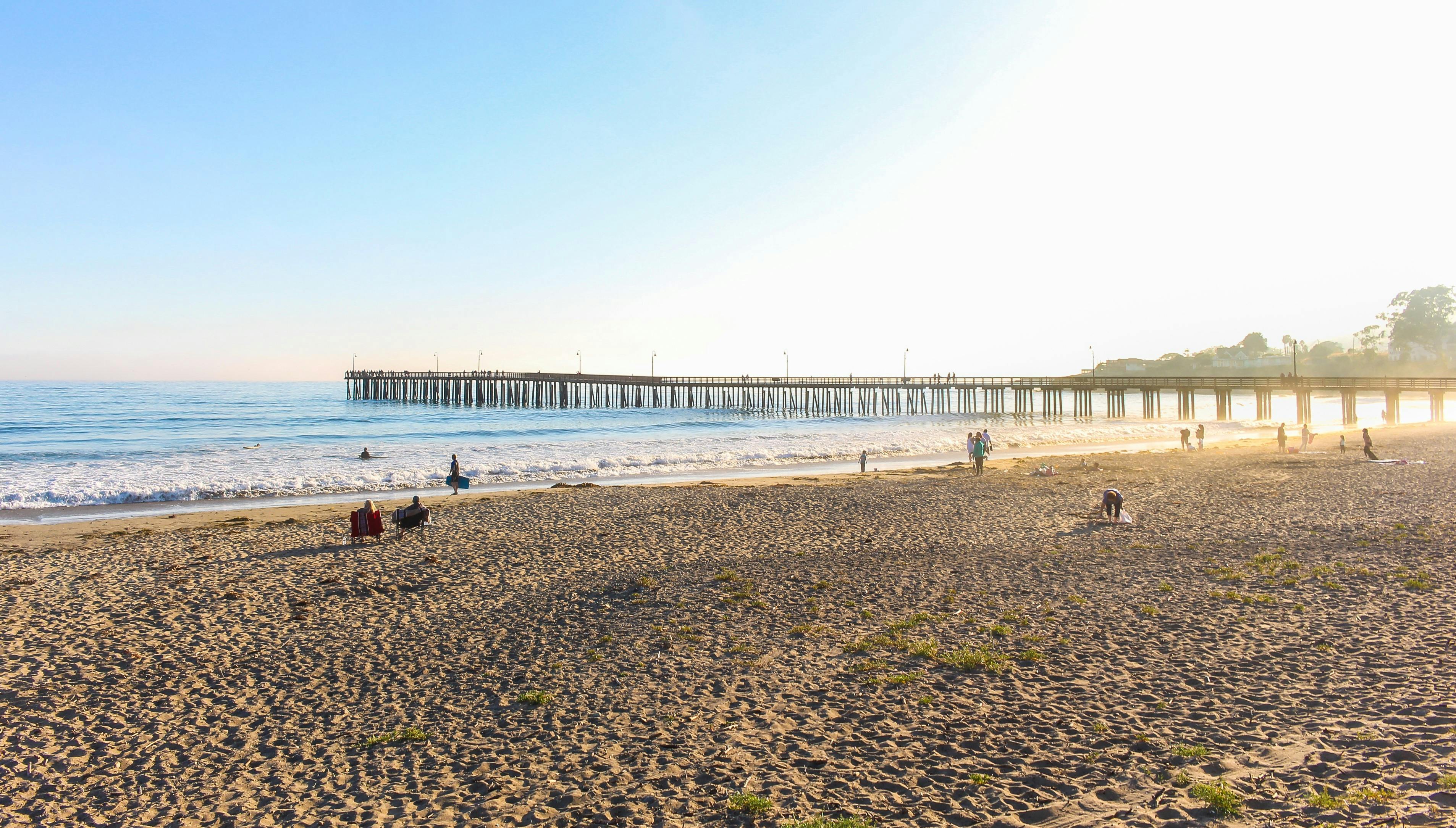 People hanging out on a sandy beach near the shore with a wooden pier out leading out into the ocean.